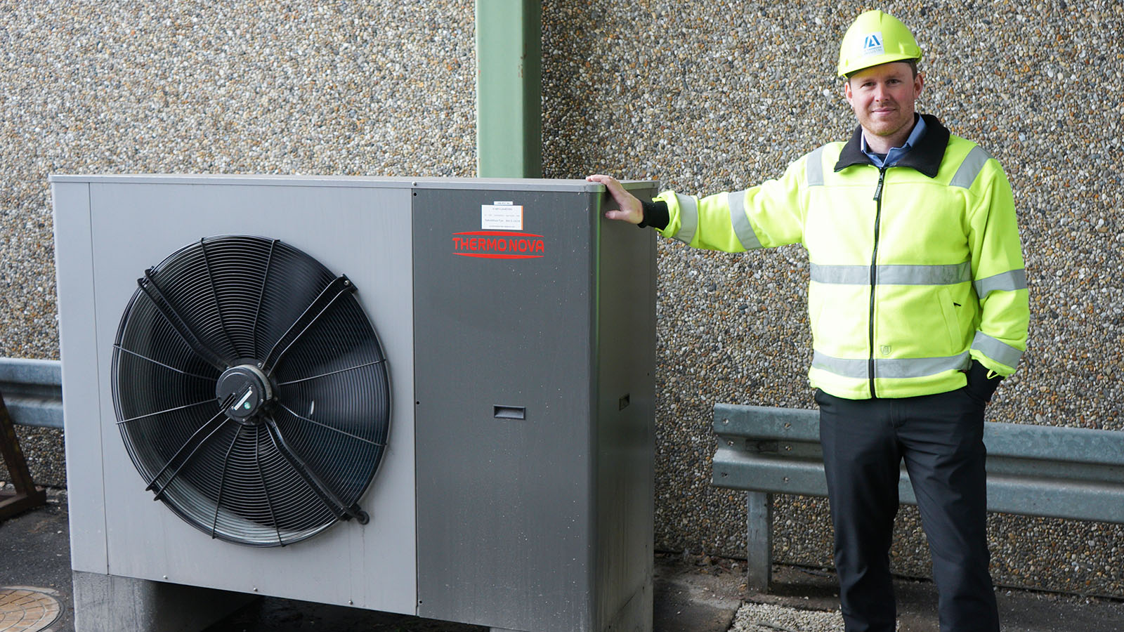 An Ib Andresen Industri employee stands beside a heat pump, casually resting a hand on it.